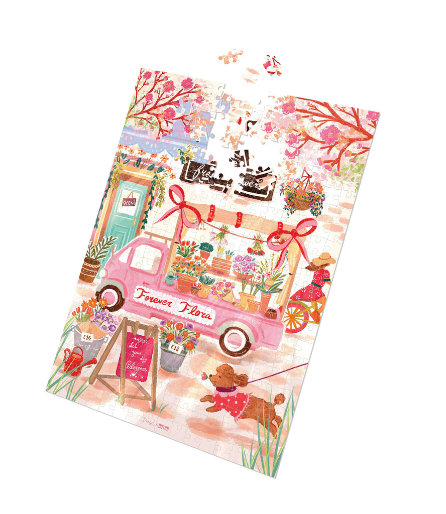 Fresh Flowers - 1000pc puzzle-Diesel & Dutch-A vintage pink flower truck overflows with vibrant blooms, creating a picturesque market scene full of charm and warmth. Nearby, a playful pup dressed in a polkadot outfit trots along, adding a whimsical touch. Surrounded by blossoming trees and a quaint little shop, this delightful puzzle captures the essence of a bright and cheerful spring day. Designing to the latest in homeware design, as well as key colours and trends, we are committed to regularly updating 