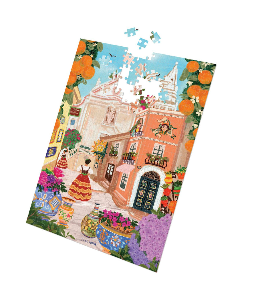 Sicilian Dreamscape - 1000pc puzzle-Diesel & Dutch-Transport yourself to the sun-kissed streets of Taormina with this vibrant art print! Let the essence of Sicily fill your space as you admire the iconic Trinacria symbol and breathe in the sweet scent of orange trees. This illustration captures the charm of Taormina's landmarks - the stunning Church of St. Joseph and the quaint clock tower. Adorned with colouful ceramic pots and the lively scenes of locals going about their day, its a true celebration of Si